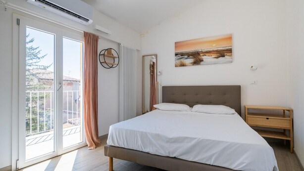 A bedroom with a bed, a wooden nightstand, a window with curtains, an air conditioning unit, and a painting on the wall.