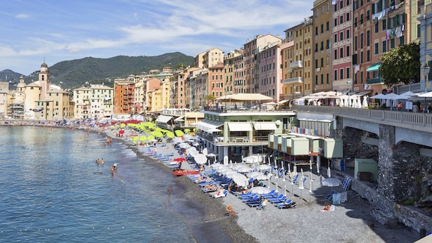 A coastal town with a beach lined with umbrellas and sun loungers. Buildings with balconies and a bridge are visible. - Nel Cuore del Borgo di Camogli by Wonderful Italy (Camogli)