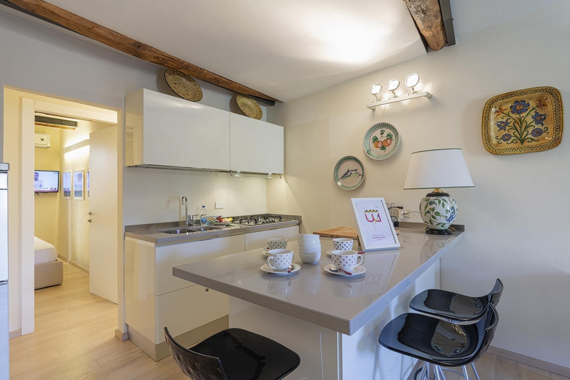 A compact kitchen with a central island, a sink, and a stove. There are two chairs at the island and a small table with a lamp.