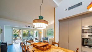 Eine moderne Küche mit eingebauten Backöfen, ein Essbereich mit einem Holztisch und Stühlen sowie ein großes Fenster mit Blick ins Freie.