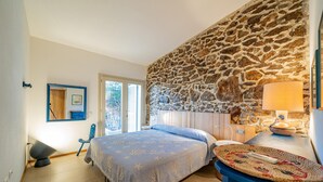 Ein Schlafzimmer mit Steinsandsteinwand, einem Bett mit blauer Bettdecke, einem Sessel, einem Nachttisch mit Lampe und einem Fenster mit Jalousien.