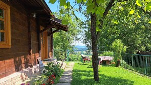 Eine Holzhütte mit Veranda, ein Garten mit bunten Blumen, einem Tisch und Stühlen sowie Blick auf die Berge im Hintergrund.