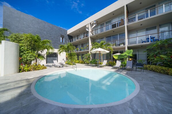 Piscina con sillas de descanso y sombrillas, rodeada de follaje y un edificio de varios pisos con balcones.