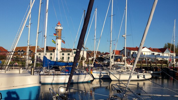 Ein Yachthafen mit mehreren Segelbooten, ein Leuchtturm und im Hintergrund Gebäude mit roten Dächern.