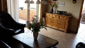 Ein Wohnzimmer mit einem Ledersofa, einem Couchtisch mit Blumen in einer Vase, einem Holzschrank mit Radio und Blick durch eine Glastür auf eine Backsteinmauer und einen Garten.