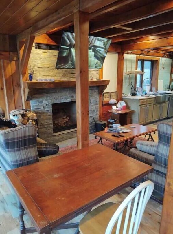 A cozy cabin interior with a stone fireplace, wooden beams, plaid furniture, and a kitchen area. - Remote private, w/5 acre pond and w/Shooting Range @ Devils Hopyard State Park (East Haddam)