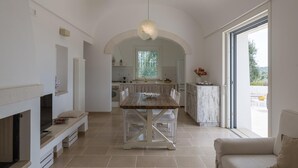 A spacious living area with a fireplace, a dining table, and a sofa. There is a kitchen in the background with a large window. - Villa Degli Ulivi con Piscina by Wonderful Italy (Ostuni)