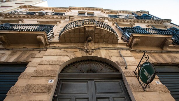 A building facade with a large, dark wooden door, a stone archway, and a metal sign hanging from the right side. - Agatea a due Passi dal Duomo by Wonderful Italy (Catania)