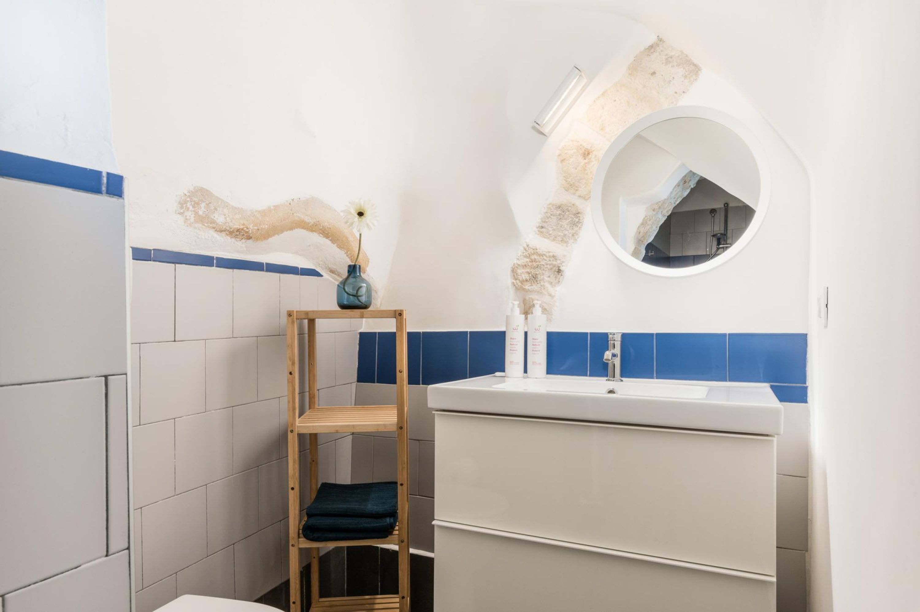 Een badkamer met een wit wastafelmeubel, blauwe tegels, een houten plank met handdoeken en een spiegel met een steenachtige textuur.