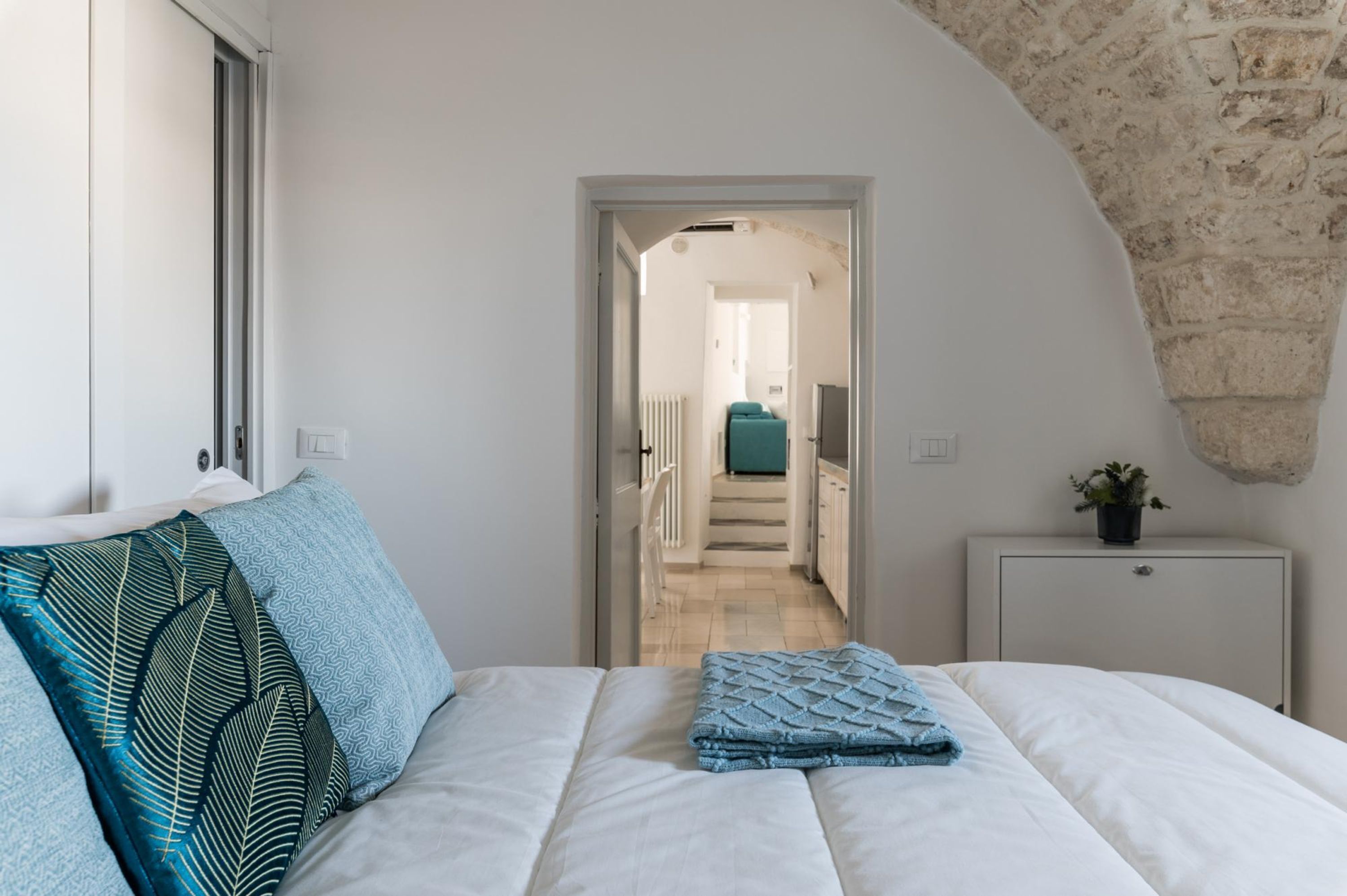 A bedroom with a bed, pillows, a bedside table, and a view of a hallway leading to a kitchen.