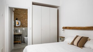 A bedroom with a bed, a wooden headboard, a brick wall, a white wardrobe, and a bathroom with a sink and mirror. - San Michele Apts by Wonderful Italy - Aluminium (Bologna)