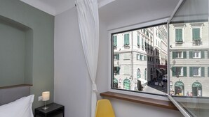 Habitación de hotel con cama, mesita de noche, lámpara, una silla amarilla y ventana con vistas a edificios.