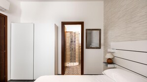 Une chambre moderne avec un lit, une table de chevet avec une lampe, une armoire et un espace douche visible à travers une porte ouverte.