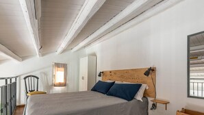 Ein Schlafzimmer mit einem Holz-Kopfteil, einem Bett mit grau-blauer Bettwäsche, einer Nachttischlampe, einem kleinen Nachttisch aus Holz und einem Spiegel an der Wand.