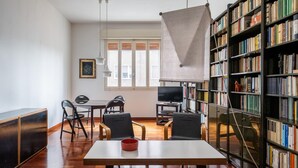 A room with a wooden floor, a white table with a red bowl, two black chairs, a black bookshelf, a television, and a window with blinds.