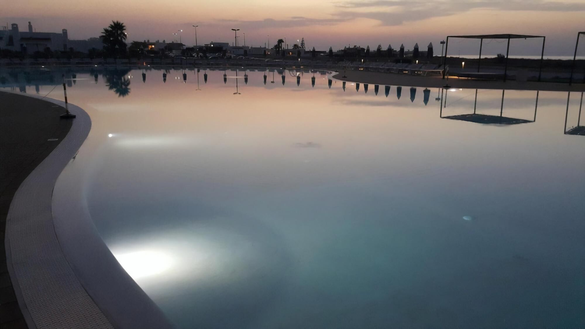 A calm swimming pool with clear reflections, surrounded by a low wall and a few street lamps.