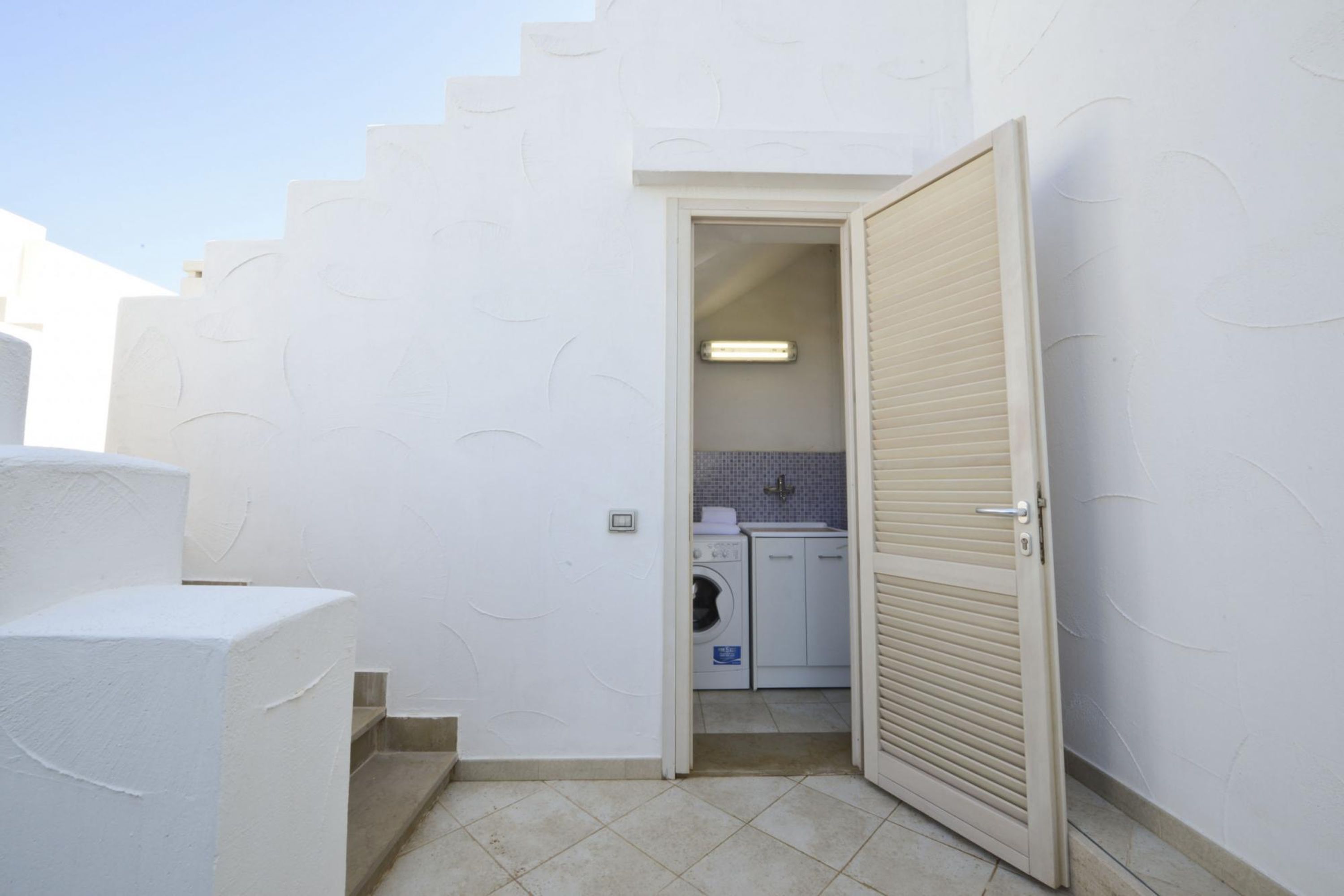 Une petite buanderie comprenant un lave-linge et un sèche-linge, un petit évier et une porte donnant sur un escalier.