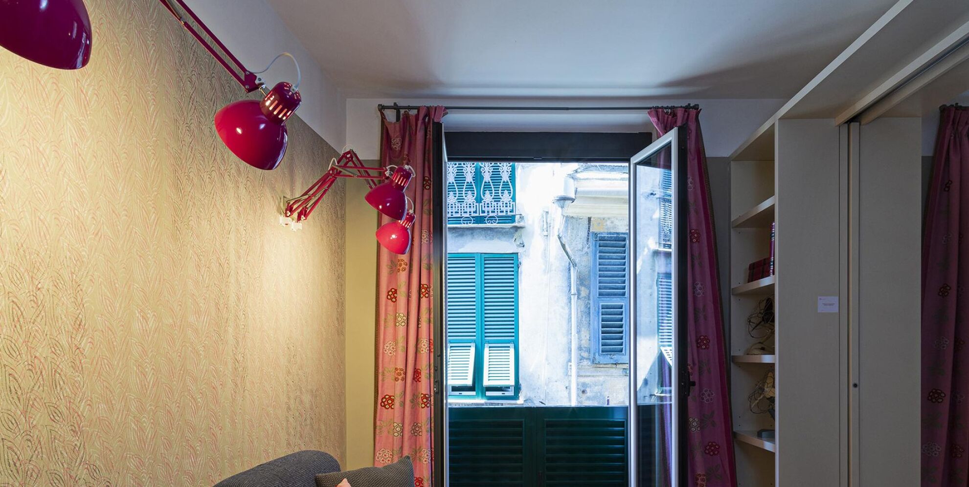 A narrow hallway with a red lamp, a door leading to a balcony, and a view of a building with blue shutters.