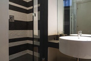 Un baño moderno con pared de azulejos a rayas negras y blancas, cabina de ducha de vidrio, lavamanos redondo con espejo y un perchero.