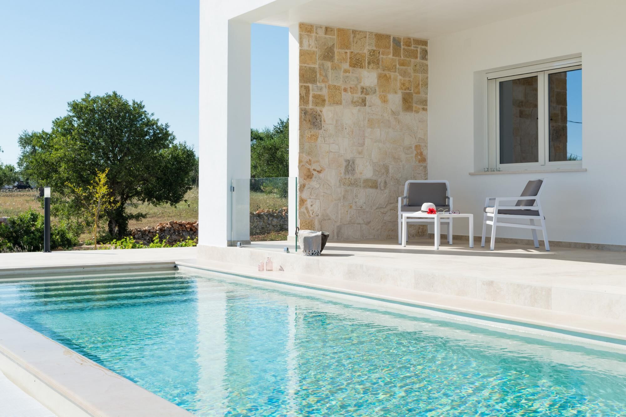 A pool area with a stone wall, two white chairs, and a table.