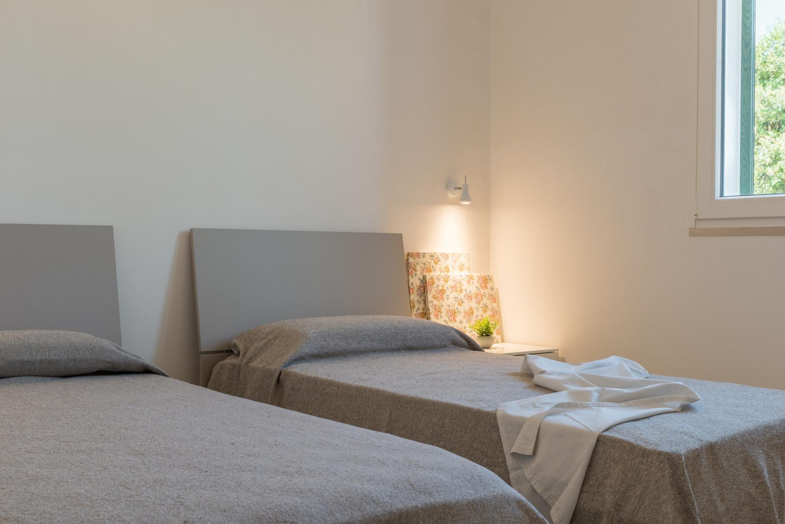 Two single beds with grey linens, a small table with a lamp, and a window with a view of greenery.