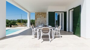 An outdoor dining area with a table set for six, surrounded by white chairs, a stone wall, and a view of a swimming pool and greenery. - Charming Villa With Pool by Wonderful Italy (Ostuni)