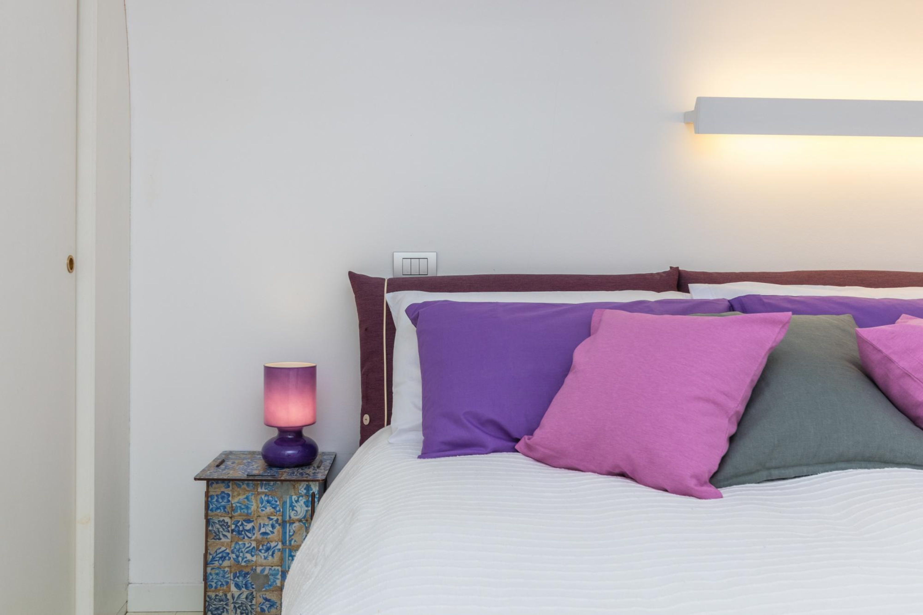 Un lit avec un matelas blanc et une tête de lit ornée d’un bordure marron. On y trouve quatre oreillers de couleurs violette, rose et verte. Une petite table avec une nappe à motifs bleus et blancs et une lampe violette se trouve à côté du lit.