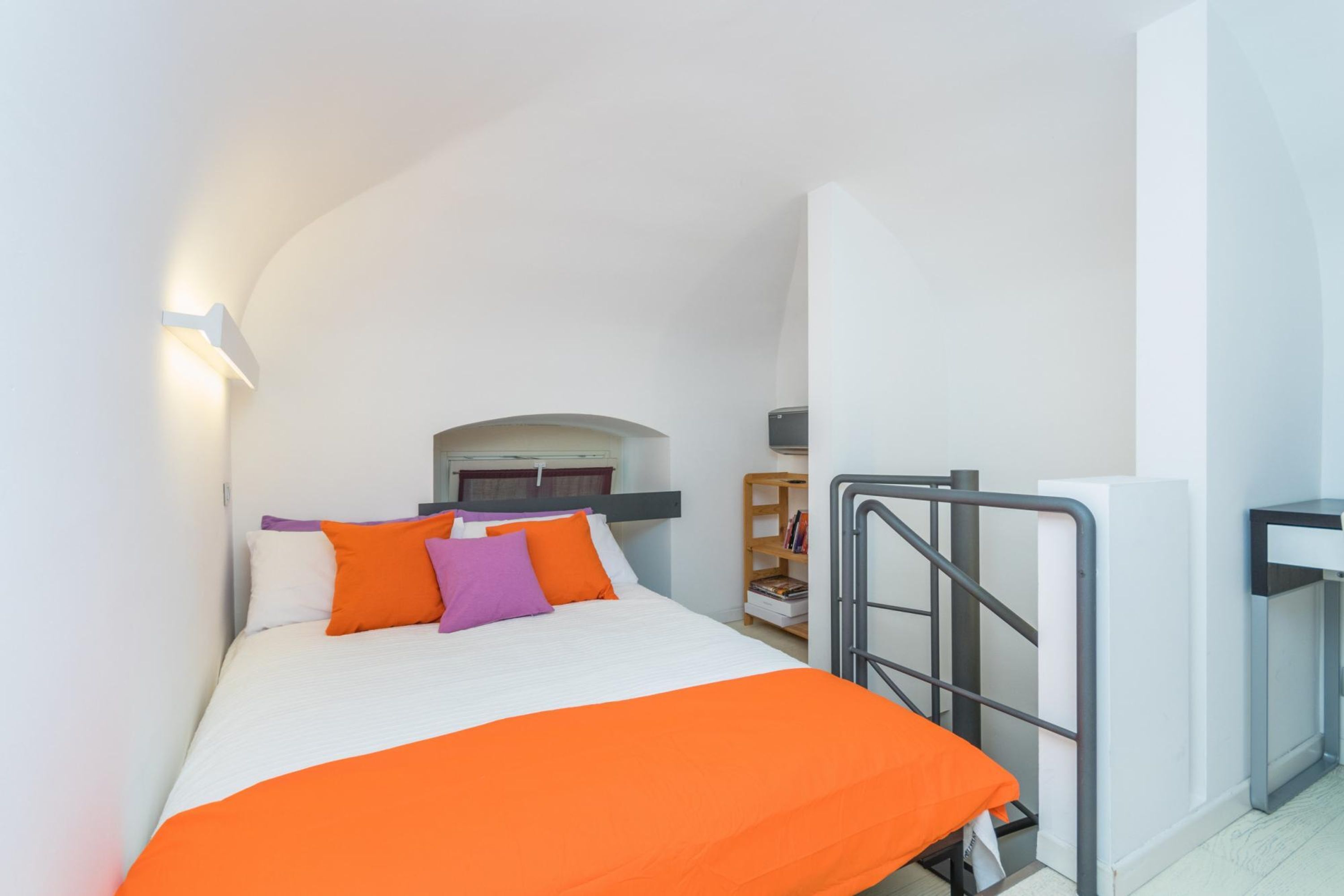 A modern hotel room with a bed, a nightstand, a small table, and a bookshelf.