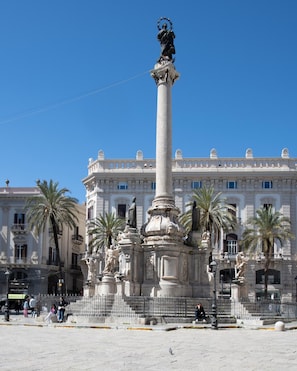 A large monument with a statue on top, surrounded by palm trees and a building with classical architecture in the background.