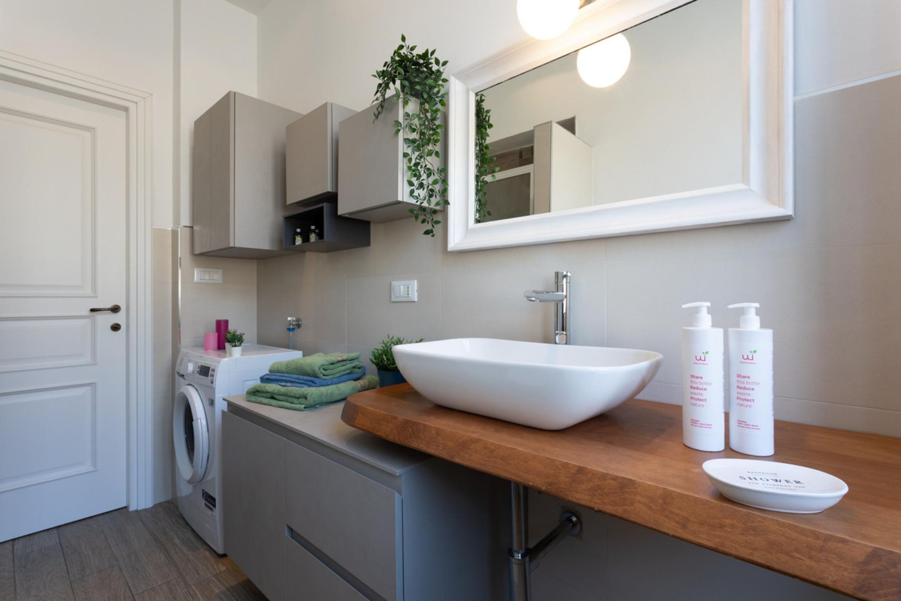 A modern bathroom with a large mirror, a white sink, and a wooden countertop. There are two bottles of shampoo and a bowl on the counter.