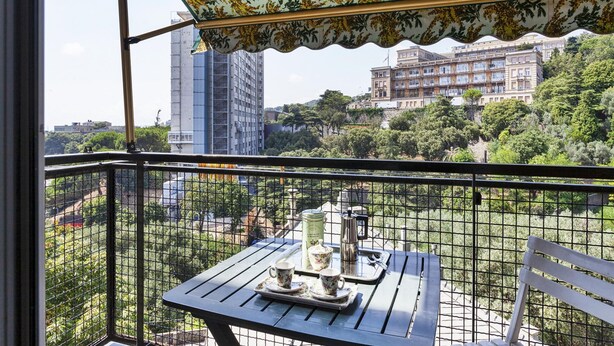 A balcony with a table set for tea, overlooking a cityscape with buildings and greenery.