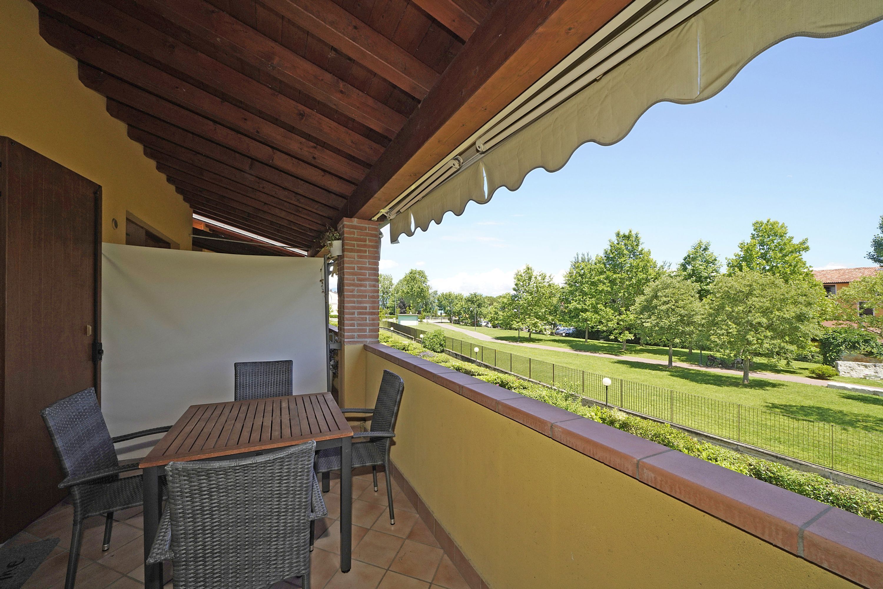 A covered patio with a wooden table and wicker chairs, overlooking a well-maintained lawn and distant buildings.