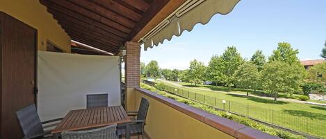 A covered patio with a wooden table and wicker chairs, overlooking a well-maintained lawn and distant buildings.