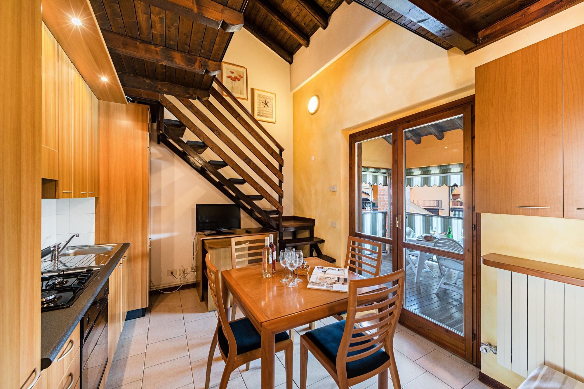Cocina compacta con armarios de madera, comedor con mesa y sillas, televisor y una escalera que sube a un nivel superior.