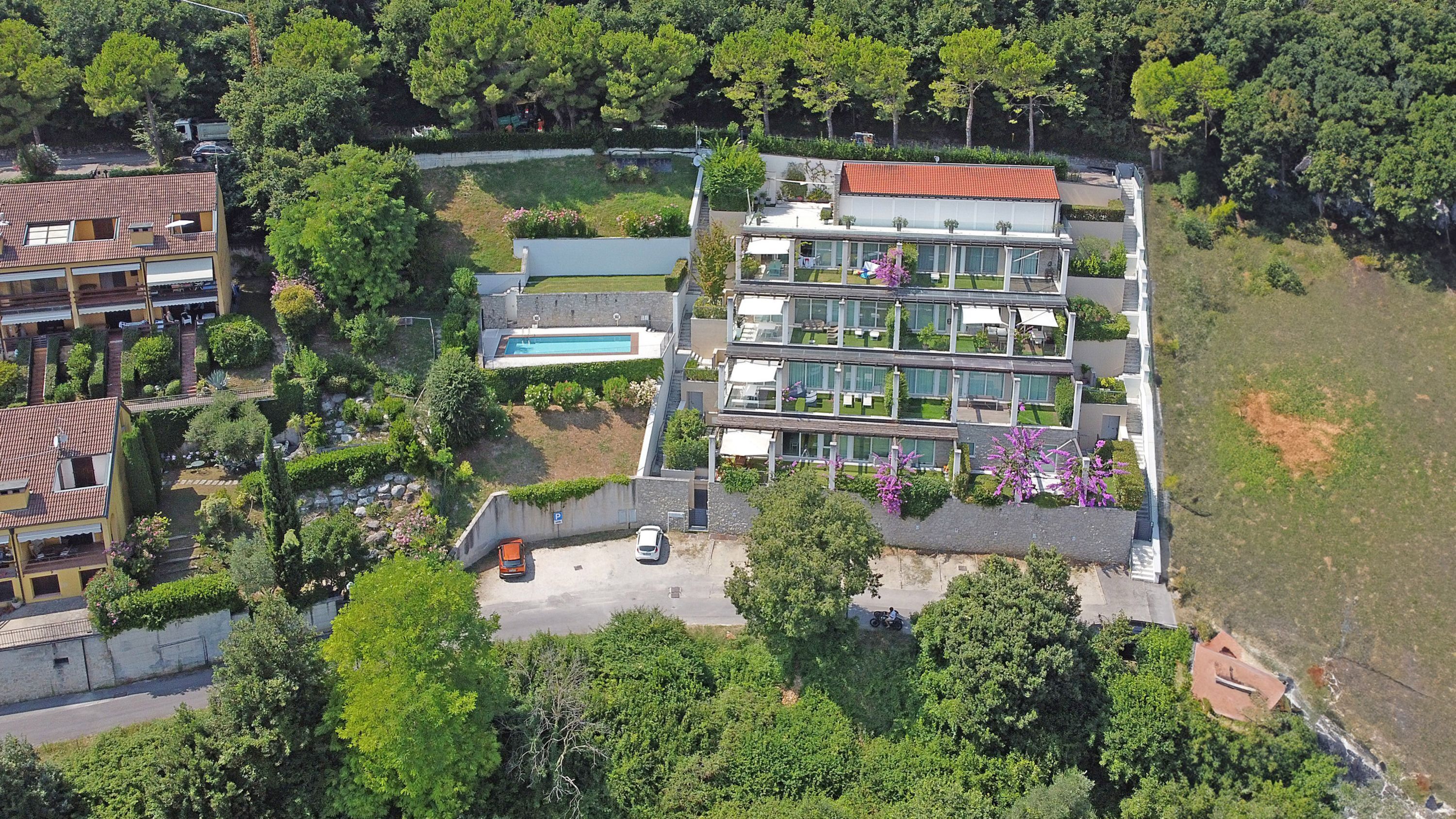 A modern multi-story residential building with a swimming pool, surrounded by lush greenery and other residential structures.
