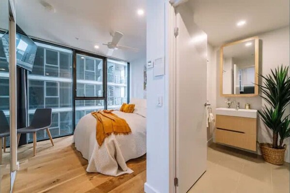 A modern bedroom with a large window, a bed with a white blanket, a chair, and a bathroom with a mirror and sink. - Pool, Free Carpark, Gym, Compact and Cosy Studio (South Brisbane)