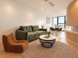 A modern living room with a green sofa, a brown chair, a round coffee table, and a dining area with a table and chairs.