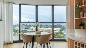 Habitación moderna con un ventanal grande que ofrece vistas a la ciudad, una mesa redonda con sillas y un aparador de madera con objetos decorativos.