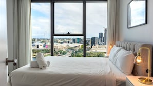 Habitación de hotel con una cama grande, una mesita de noche, una lámpara y vistas panorámicas de la ciudad a través de amplias ventanas.