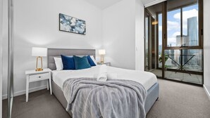 Une chambre d’hôtel moderne avec un grand lit, deux tables de chevet avec des lampes, une vue sur la ville à travers de grandes fenêtres et un tableau au mur.
