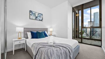 A modern hotel room with a large bed, two bedside tables with lamps, a city view through large windows, and a painting on the wall.