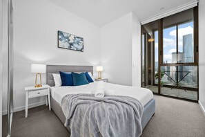 Una habitación de hotel moderna con una cama grande, dos mesitas de noche con lámparas, vistas a la ciudad a través de amplios ventanales y un cuadro en la pared.