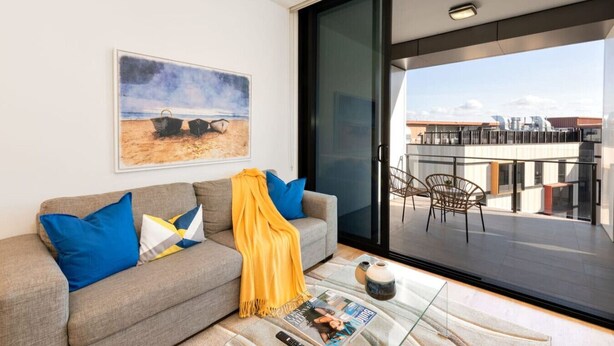 Un salón moderno con un sofá gris, cojines azules y amarillos, un cuadro de barcos en la playa y un balcón con mobiliario exterior.