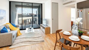 Una sala moderna con sofá, una mesa de centro y una zona de comedor con mesa para dos. Hay una puerta corrediza de vidrio que da a un balcón con mobiliario exterior.