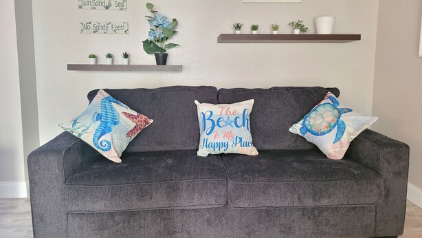 A grey sofa with three decorative pillows, a wall-mounted shelf with plants, and a framed picture. - On The Sand - Oceanfront - Dream Location (Huntington Beach)