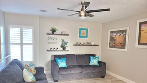 A living room with a gray sofa, a ceiling fan, wooden flooring, and wall-mounted shelves with plants and decorative items. - On The Sand - Oceanfront - Dream Location (Huntington Beach)
