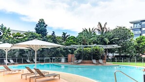 Une piscine entourée de chaises longues et de parasols, avec de la verdure tout autour et un bâtiment en arrière-plan.