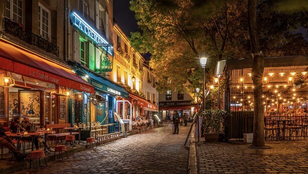 A cobblestone street lined with restaurants and cafes, featuring outdoor seating and illuminated signage. - New architect's'apartment located in the heart of the village of Montmartre (Paris)