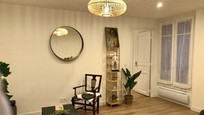 A room with a wooden floor, a round mirror, a wooden chair, a black table with a plant, a tall wooden bookshelf, and a window with curtains. - New architect's'apartment located in the heart of the village of Montmartre (Paris)