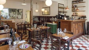 A restaurant interior with wooden tables and chairs, hanging pendant lights, a bar area with shelves of bottles, and a checkered floor. - New architect's'apartment located in the heart of the village of Montmartre (Paris)